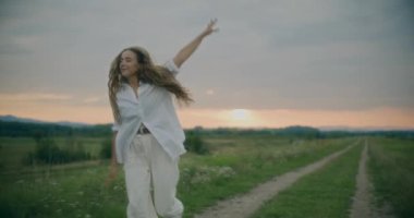 Yavaş çekim kaygısız genç kadın gün batımında çayırda gökyüzüne karşı koşarken ve dönerken.