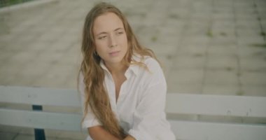 Ağır çekimde, sarı saçlı depresif genç bir kadın bankta otururken gözlerini kaçırıyor.