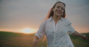 Gün batımında gökyüzüne karşı çayırda eğlenen mutlu genç kadının yavaş çekim görüntüleri.