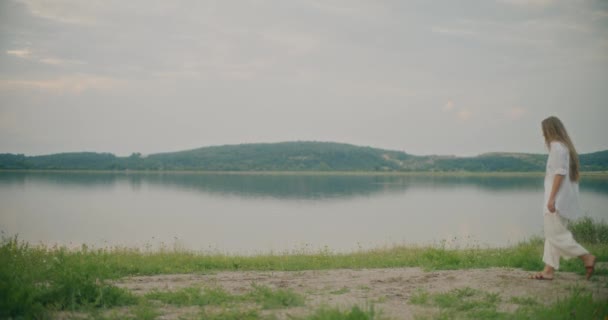 一个疲惫的金发女人在湖畔的天空中缓缓地走着 站在山前的湖畔 — 图库视频影像