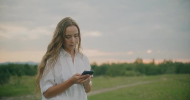 在草地上散步的金发美女通过智能手机发短信的慢镜头 — 图库视频影像