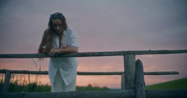 Gün batımında köprüye yaslanmış düşünceli genç bir kadının yavaş çekim sahnesi.