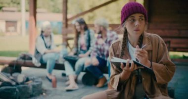 Düşünceli genç bir kadın arka planda oturan arkadaşlarıyla günlüğe yazıyor.