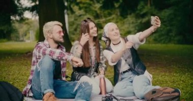 Mutlu genç bir kadın arkadaşlarıyla parkta çimlerde otururken selfie çekiyor.