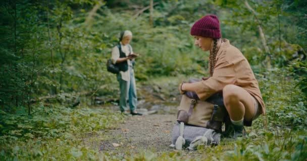 森の植物の近くで曲がっている間編まれた帽子のパッキングのバックパックを身に着けている女性 — ストック動画