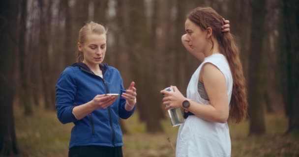 スマートフォンを使用して 公園に立っている間に女性の友人と話す若いスポーツウーマン — ストック動画