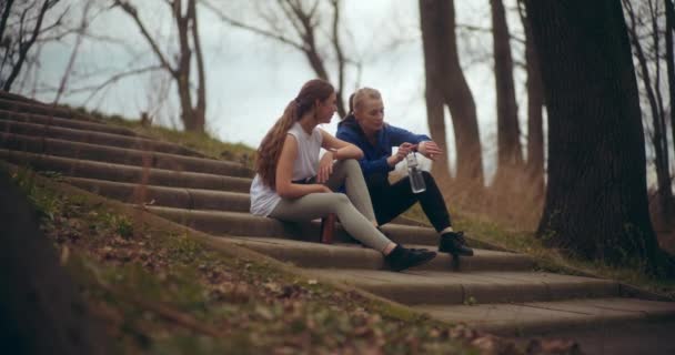 若い女性は腕時計をチェックして 公園のステップに女性の友人と座って — ストック動画