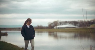 Elleri ceplerinde genç bir kadın kaşif tatil sırasında göl kenarında bulutlu gökyüzüne doğru yürüyor.