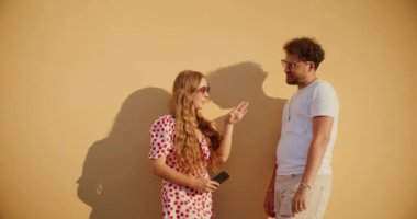 Sarışın genç kadın güneş gözlüğü takıyor ve erkek arkadaşıyla duvara karşı konuşuyor.