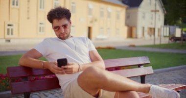 Parktaki bankta otururken güneş gözlüğü takan genç adam cep telefonu kullanıyor.
