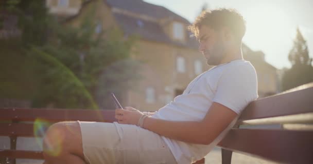 年轻人坐在公园的长椅上 一边用智能手机发短信 — 图库视频影像