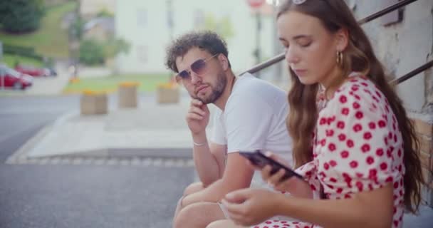 スマートフォンを使用している若い女性は 徒歩道で周りを見回しながらボーイフレンドに座って — ストック動画
