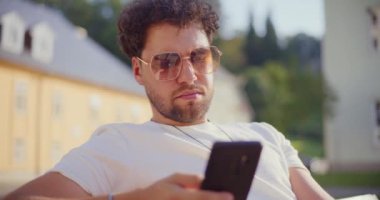 Güneş gözlüğü takan genç adam parkta otururken cep telefonundan mesaj atıyor.