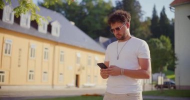 Hafta sonu bahçede yürürken güneş gözlüğü takan genç adam akıllı telefon kullanıyor.