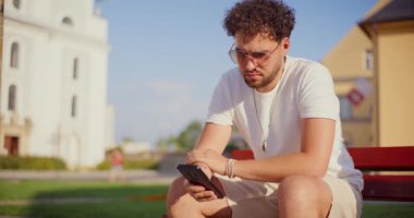 Güneşli bir günde bahçede bankta otururken cep telefonu kullanan yakışıklı adam.