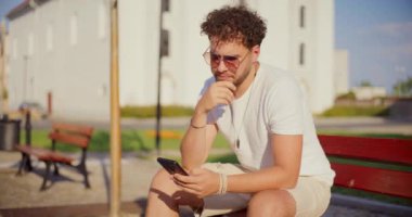 Genç adam güneşli bir günde parkta bankta otururken akıllı telefon kullanıyor.