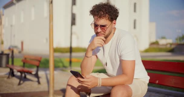 Молодой Человек Использует Смартфон Сидя Скамейке Парке Солнечный День — стоковое видео