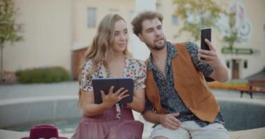 Gülümseyen adam cep telefonuyla selfie çekiyor. Elinde dijital tabletle bankta oturuyor.