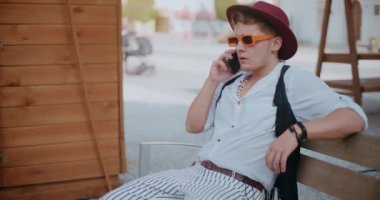Güneş gözlüklü ve şapkalı genç adam bankta otururken cep telefonuyla konuşuyor.