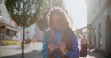 Çekici genç bir kadın güneşli bir günde yaya yolunda yürürken akıllı telefondan mesaj atıyor.