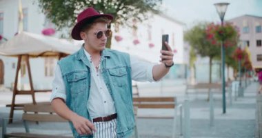 Moda kıyafetleri içinde yakışıklı bir adam yürüme yolunda akıllı telefondan selfie çekiyor.
