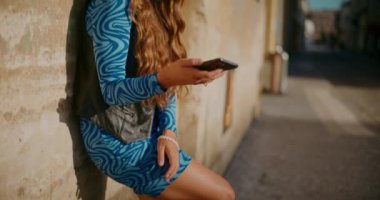 Sarışın genç bir kadın, yaya yolunda duvara yaslanırken cep telefonundan mesaj atıyor.