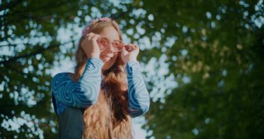 Gülümseyen sarışın kadın güneş gözlüğü takıyor ve bahçede dikilirken etrafına bakıyor.