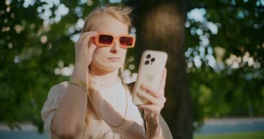 Sarışın genç kadın güneş gözlüğünü çıkarıyor ve güneşli bir günde parkta cep telefonu kullanıyor.