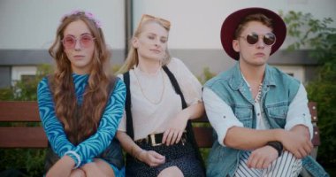 Parkta güneş gözlüklü, erkek ve kadın arkadaşlarının arasında oturan gülümseyen genç kadın.