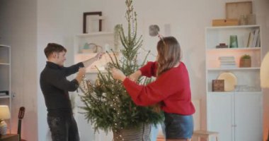 Genç adam ve kadınlar oturma odasında ışıklandırılmış ışıklarla Noel ağacını süslüyorlar.