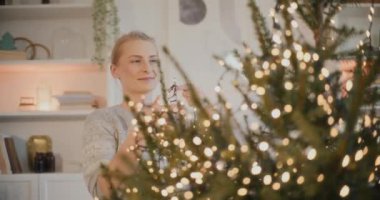Gülümseyen bir kadın Noel ağacını süslerken evinde parıldayan ışıklarla