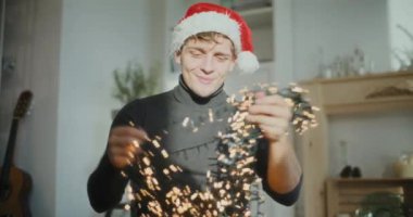 Noel Baba şapkası takan genç adam evde Noel süsü olarak parlayan ışıkları sıralıyor.