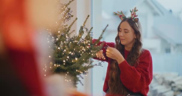 Jonge Vrouw Versieren Boom Met Gloeiende Lichten Thuis Tijdens Kerstvakantie — Stockvideo