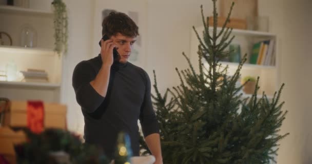 Forstyrret Ung Mand Taler Mobiltelefon Mens Stående Ved Juletræ Derhjemme – Stock-video