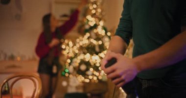 Oturma odasındaki bayan arkadaşın Noel ağacına kurşunlu ışık bağlayan adam.