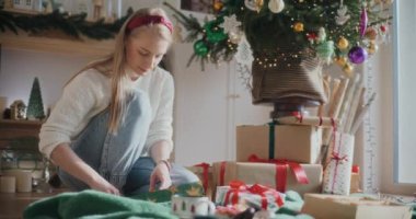 Güzel genç bir kadın, süslenmiş Noel ağacının yanında otururken hediyeleri paketliyor.