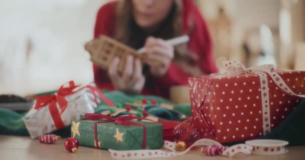 自宅の背景にダンボールハウスの装飾を着色する女性と床に包まれたクリスマスプレゼント — ストック動画