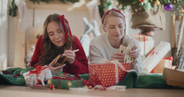 圣诞节期间 漂亮的妹妹们在家里躺在地板上做纸制房屋装饰品 — 图库视频影像