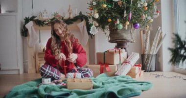 Mutlu genç bir kadın, evde battaniyeye oturmuş, Noel hediyesine kurdele bağlıyor.