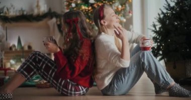 Kahve fincanı taşıyan neşeli genç kız kardeşler Noel 'de evde sırt sırta otururken şarkı söylüyorlar.