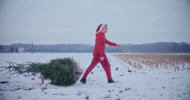 身穿红色保暖衣服 头戴圣诞礼帽 拖着雪地上挂着圣诞树的雪橇的人笑了 — 图库视频影像