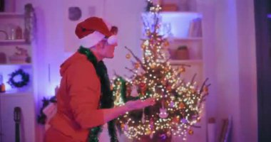 Noel Baba şapkalı ve güneş gözlüklü genç adam. Omuzlarında yeşil süslemelerle aydınlatılmış evde Noel boyunca dans ediyor.
