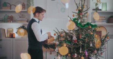 İyi giyimli genç bir adamın evdeki süslü Noel ağacına takılıp süslenmesinin yan görüntüsü.
