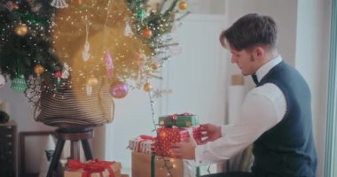 İyi giyimli bir gencin Noel ağacının yanında hediye kutuları düzenleyişinin yan görüntüsü.
