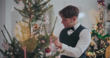 İyi giyimli smokinli genç adam Noel ağacını evde süslerle süslüyor.