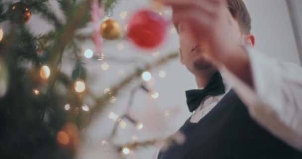 自宅でクリスマスツリーに赤い問題を掛けている若者の低い角度のビュー — ストック動画