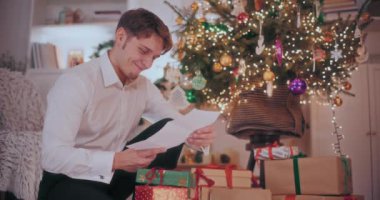 Noel hediyesini elinde tutan mutlu genç adam süslü evde mektup okuyor.