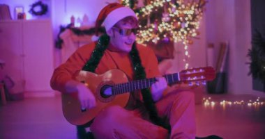 Noel Baba şapkalı ve güneş gözlüklü mutlu genç adam Noel 'de aydınlatılmış evde otururken gitar çalıyor.