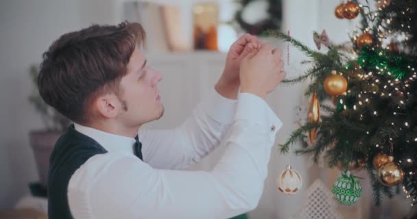 自宅で装飾されたクリスマスツリーにバブルを掛けるタキシードでよく服を着た若者の側面のビュー — ストック動画