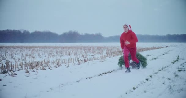 身穿红色保暖衣服头戴圣诞帽的年轻人一边跑着一边在雪地上跑着 拉着圣诞树 — 图库视频影像